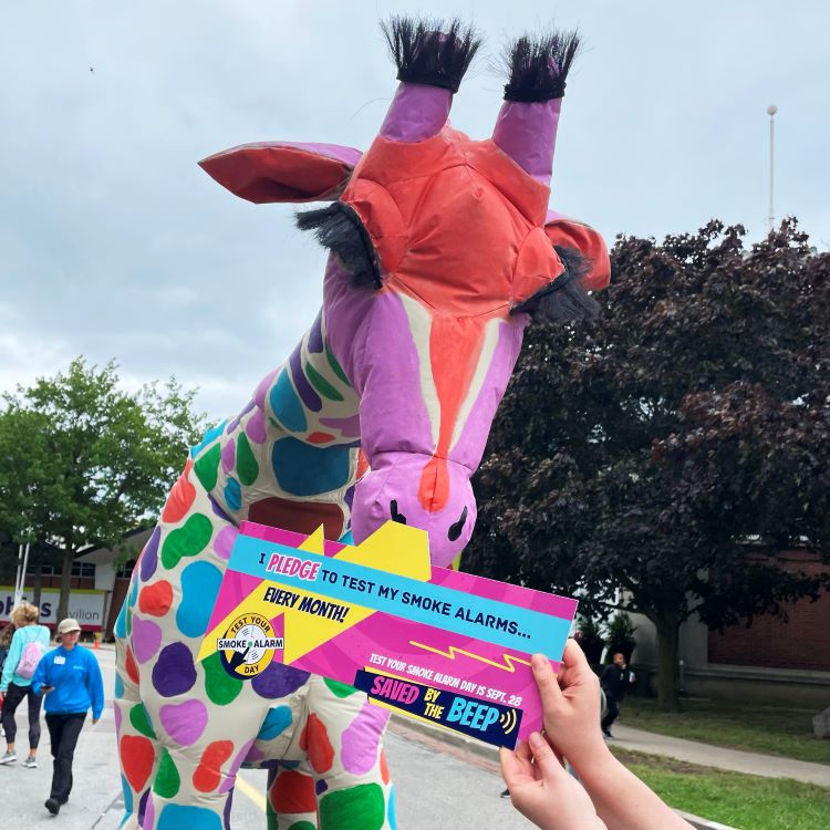 Picture of colourful Giraffe puppet at the Canadian National Exhibition with sign made by Office of the Fire Marshal, promoting this year’s Saved By the Beep fire safety campaign.