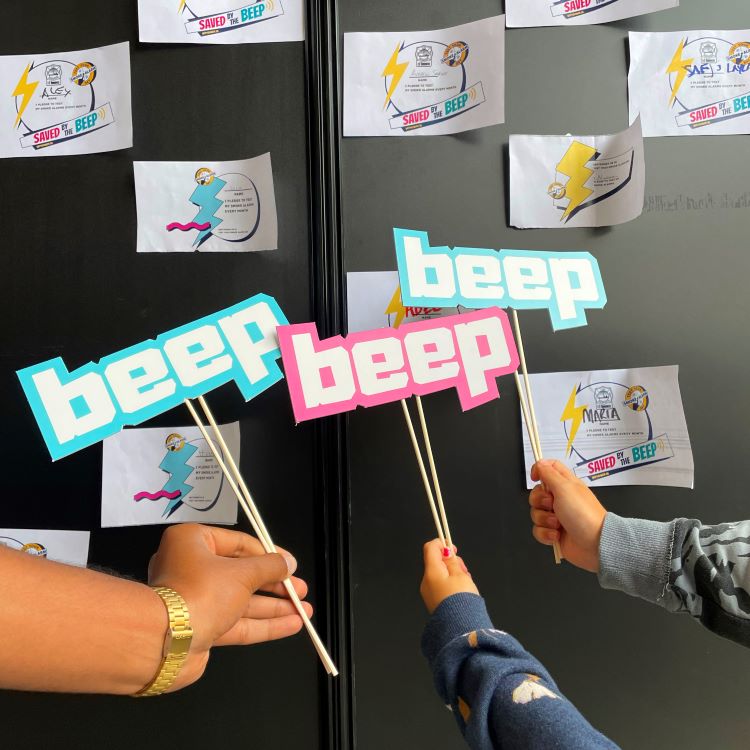 Picture of people and children holding WordArt sign with the word ‘beep’, against a pledge wall at the Canadian National Exhibition within the Toronto Fire hall as part of the Saved By the Beep fire safety campaign.